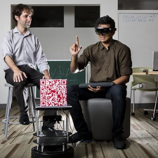 Two students talk in a classroom while one wears virtual reality glasses.
